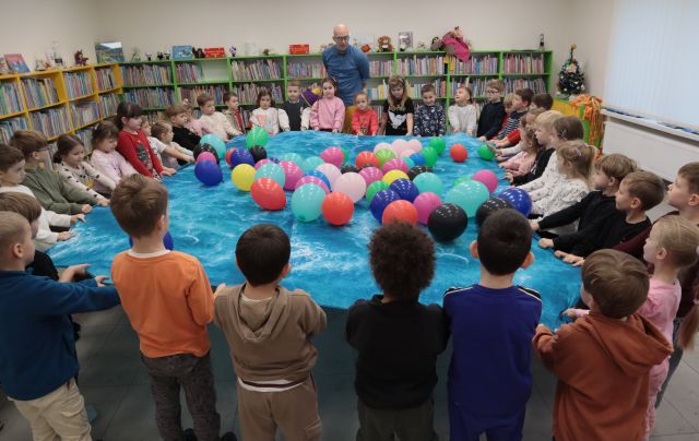 Na zdjęciu znajdują się przedszkolaki, trzymające niebieską chustę, na której znajdują się kolorowe balony. Za nimi stoi mężczyzna, w niebieskiej koszuli. W tle znajdują się półki z książkami.