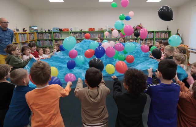 Na zdjęciu znajdują się dzieci trzymające niebieską chustę i podrzucają znajdujące się na niej balony. W tle widać półki z książkami, oraz mężczyznę po lewej stronie.