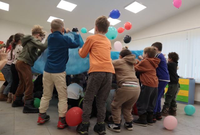 Na zdjęciu znajdują się dzieci trzymające niebieską chustę i podrzucające znajdujące się na niej balony, pod chustą znajdują się dzieci.