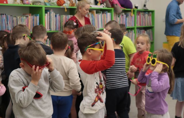 Na zdjęciu widać dzieci tyłem, które wybierają okulary ze stolika i przymierzające je. W tle widać półki z książkami