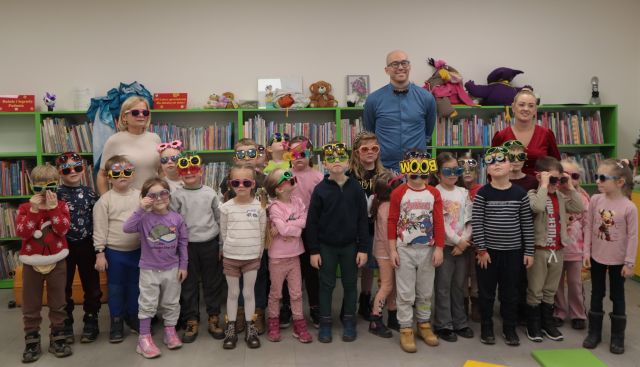 Na zdjęciu znajdują się dzieci w okularach, za nimi stoją dwie kobiety, oraz jeden mężczyzna. W tle znajdują się półki z książkami.