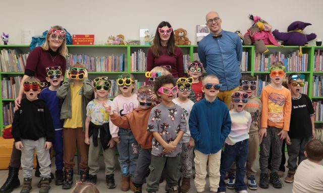 Na zdjęciu znajdują się dzieci w kolorowych okularach, za nimi stoją dwie kobiety, oraz jeden mężczyzna. W tle znajdują się półki z ksiązkami.