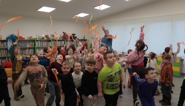 Na zdjęciu znajdują się dzieci podrzucające do góry pomarańczowe wstążki. W tle znajduję się mężczyzna, a za nim po prawej stronie okno, a po lewej półki z książkami.