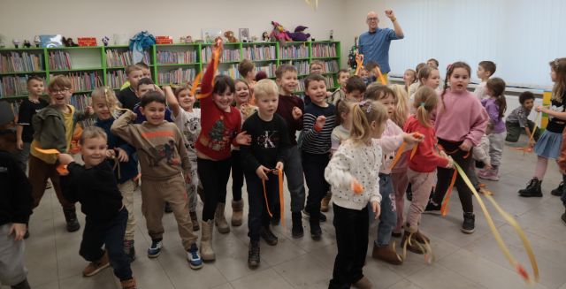 Na zdjęciu znajdują się dzieci, rzucające przed siebie wstążki. W tle znajduję się mężczyzna w niebieskiej koszuli, oraz półki z książkami.