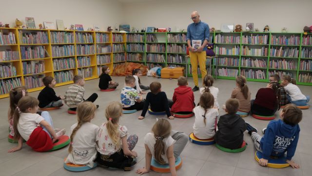 Na zdjęciu znajdują się siedzące na podłodze dzieci oraz mężczyzna stojący z książką. W tle znajdują się półki z książkami.