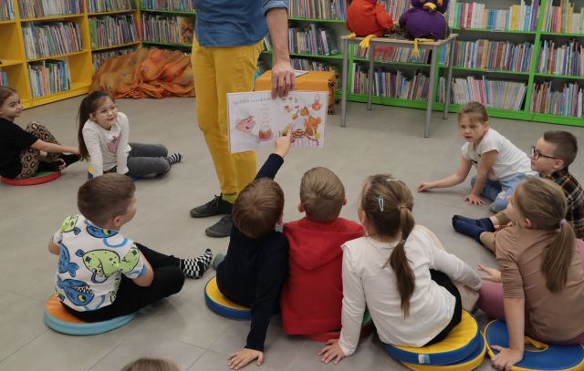 Na zdjęciu znajdują się nogi mężczyzny oraz otwarta książka, którą pokazuje dzieciom siedzącym na podłodze. W tle znajdują się półki z książkami.