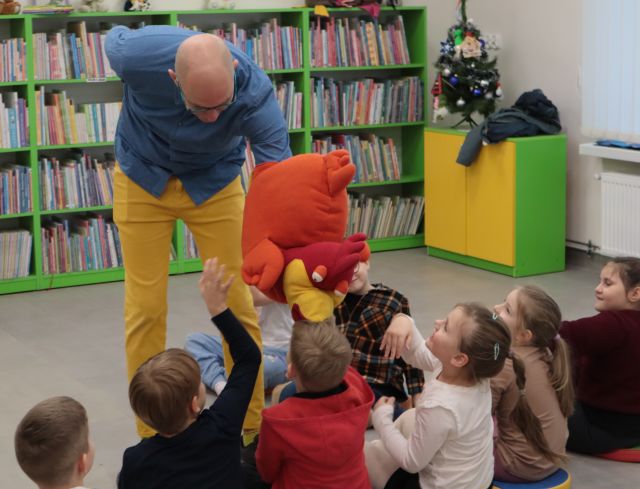 Na zdjęciu znajduje się mężczyzna pochylający się nad dziećmi z pluszową maskotką kury na ręce. W tle znajdują się półki z książkami.