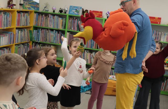 Na zdjęciu znajdują się przedszkolaki z wystawionymi rękami, w których trzymają małe pluszowe owoce i warzywa, którymi karmią maskotkę kury trzymaną na ręce przez mężczyznę, który znajduję się po prawej stronie. W tle znajdują się półki z książkami.