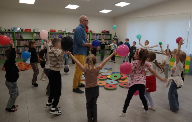 Na zdjęciu znajdują się przedszkolaki stojące w kole i wykonujące pajacyki trzymając w ręce balon. W kole znajduje się mężczyzna z balonem oraz okrągłe pufy. W tle znajdują się półki z książkami a po prawej stronie okno.