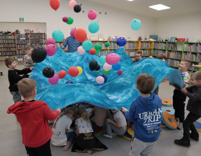 Na zdjęciu znajdują się przedszkolaki trzymające niebieską chustę i podrzucające znajdujące się na niej kolorowe balony, pod chustą znajduję się część przedszkolaków, a w tle widać półki z książkami.