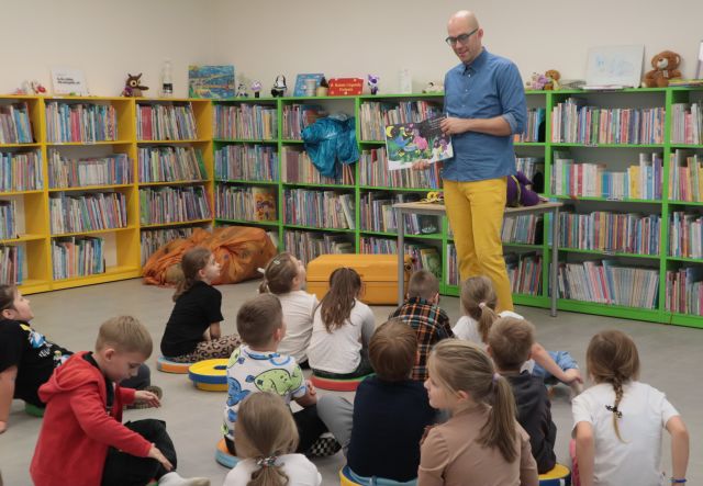 Na zdjęciu znajduję się mężczyzna z otwartą książką, którą pokazuje siedzącym na przeciwko niego przedszkolakom. W tle znajdują się półki z książkami.