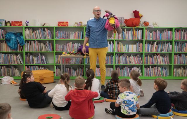 Na zdjęciu znajduje się mężczyzna z dużą fioletową maskotką kaczki na ręce, na przeciwko niego siedzą dzieci w wieku przedszkolnym. W tle znajdują się półki z książkami.