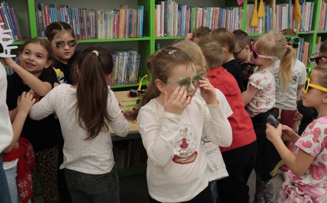 Na zdjęciu znajduję się dziewczynka poprawiająca okulary, a za nią znajdują się dzieci wybierające okulary ze stolika, który stoi na tle półek z książami.