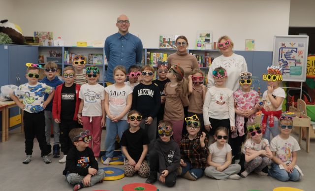 Na zdjęciu znajdują się przedszkolaki część z nich siedzi a część stoi. Za nimi stoi mężczyzna oraz dwie kobiety. W tle znajdują się półki z grami i szafa.