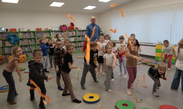 Na zdjęciu znajdują się dzieci rzucające przed siebie pomarańczowe wstążki, za nimi stoi mężczyzna. W tle znajdują się półki z książkami i okno po prawej stronie.