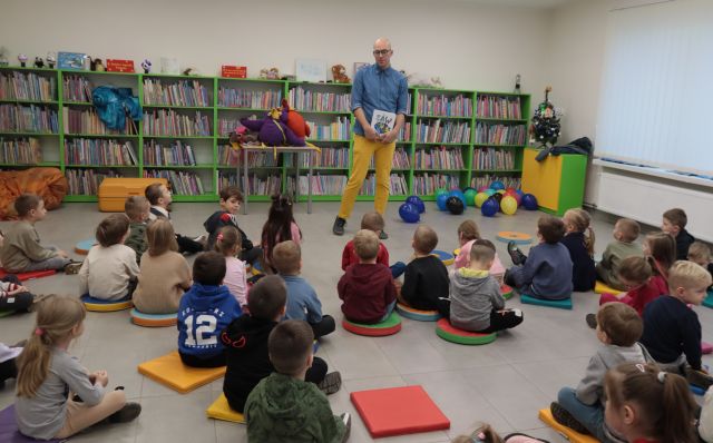 Na zdjęciu znajduję się mężczyzna trzymający książkę, przed nim siedzą dzieci w wieku przedszkolnym. W tle znajdują się półki z książkami oraz okno po prawej stronie.