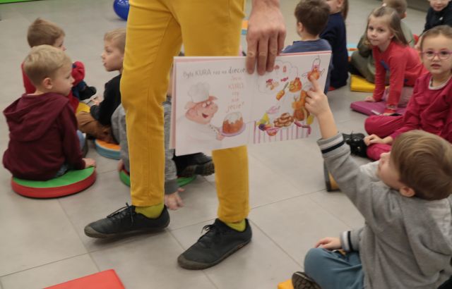 Na zdjęciu znajdują się nogi mężćzyzny, który trzyma otwartą książkę pokazując ją siedzącym dzieciom, jeden chłopiec wskazuje na ilustrację palcem.