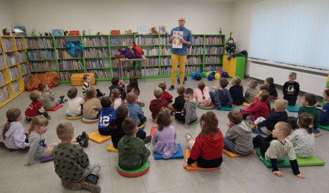 Na zdjęciu znajduję się mężczyzna z otworzoną książką, przed nim siedzą dzieci w wieku przedszkolnym. W tle znajdują się półki z książkami. 