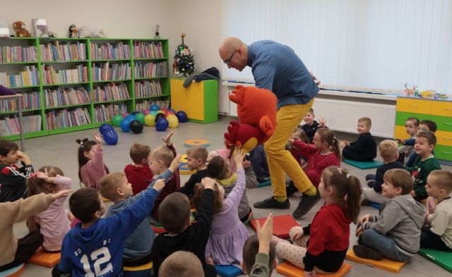 Na zdjęciu znajduję się mężczyzna pochylający się z pomarańczową maskotką kury nad siedzącymi na podłodze przedszkolakami, które wyciągają ręce w stronę maskotki. W tle znajdują się okna, półki z książkami oraz balony.