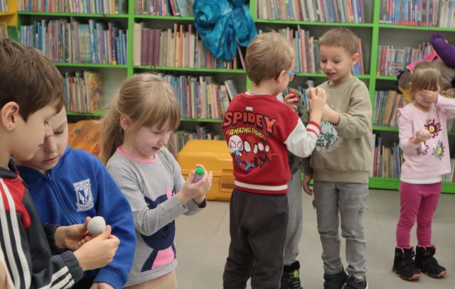 Na zdjęciu znajdują się szóstka dzieci, która ogląda trzymające w dłoniach małe zabawkowe owoce lub warzywa, dwóch chłopców stoi na przeciwko siebie i pokazują je sobie pokazują. W tle znajdują się półki z książkami.