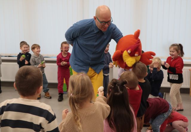 Na zdjęciu znajdują się przedszkolaki stojące w półokręgu oraz mężczyzna z pomarańczową duża maskotką kury, która zjada trzymane przez dzieci owoce i warzywa. W tle znajdują się okna.