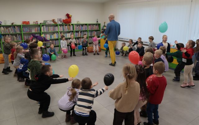 Na zdjęciu znajdują się dzieci w wieku przedszkolnym robiące przysiady w okręgu i trzymają w ręce balony. W środku koła chodzi mężczyzna. W tle znajdują się półki z książkami a po prawej stronie okna.
