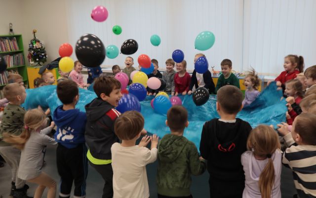 Na zdjęciu znajdują się uśmiechnięte przedszkolaki trzymające w rękach niebieską chustę i podrzucające znajdujące się na niej balony. W tle znajdują się okna.