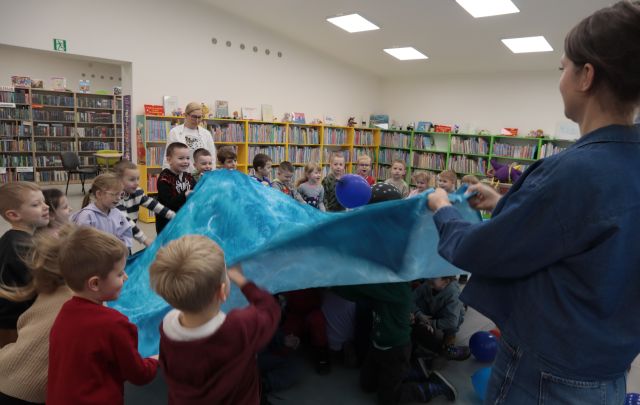 Na zdjęciu znajduje się kobieta oraz dzieci ruszające niebieską chustą, na które znajdują się dwa balony a pod chustą znajduję się reszta grupy. W tle widać kobietę oraz półki z książkami.