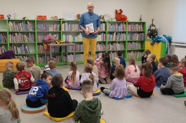 Na zdjęciu znajduję się mężczyzna stojący na wprost siedzących na podłodze dzieci i pokazuje im książkę. W tle znajdują się półki z książkami oraz stół.