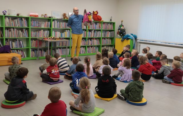 Na zdjęciu znajduję się mężczyzna stojący z maskotką kury na ręce, na przeciw niego siedzą przedszkolaki. W tle znajdują się półki z książkami, stół i szafka.