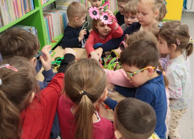Na zdjęciu znajdują się dzieci wybierające okulary ze stolika, który znajduję się przy półce z książkami.