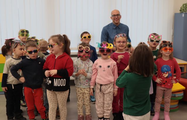 Na zdjęciu znajduje się około szesnaście dzieci, jeden chłopiec stoi tyłem. Przedszkolaki mają na sobie kolorowe okulary, za nimi stoi mężczyzna. W tle znajdują się okna.