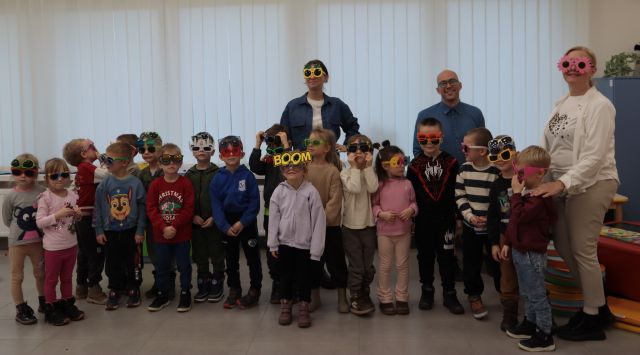 Na zdjęciu znajduję się około osiemnaście przedszkolaków, za nimi na środku i po prawej stronie stoi kobieta, a pomiędzy nimi mężczyzna. Wszyscy mają na sobie kolorowe okulary i znajdują się na tle okien.