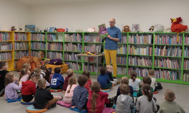 Na zdjęciu znajduję się mężczyzna pokazujący siedzącym na przeciw niego dzieciom otwartą książkę. W tle znajdują się półki z książkami.