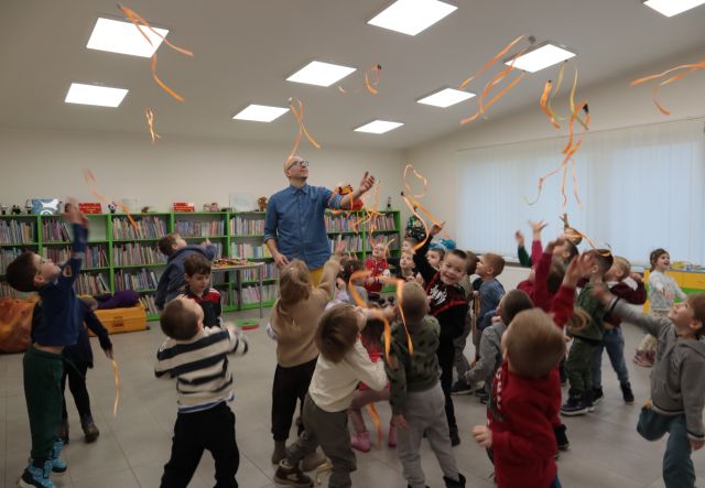 Na zdjęciu znajdują się przedszkolaki oraz mężczyzna, wszyscy rzucają w górę pomarańczowe wstążki. W tle znajdują się półki z książkami i okno po prawej stronie.