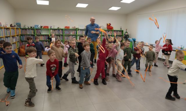 Na zdjęciu znajdują się przedszkolaki oraz stojący za nimi mężczyzna i rzucają przed siebie pomarańczowe wstążki. W tle znajdują się półki z książkami oraz okno po prawej stronie.