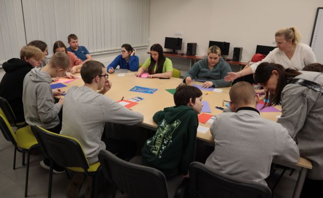 Na zdjęciu znajduję się młodzież siedząca przy złączonych stołach, na których znajdują się kolorowe kartki. Po prawej stronie kobieta w białej bluzce nachyla się z wyciągniętą ręką w stronę stołu. Znajdują się w sali komputerowej.