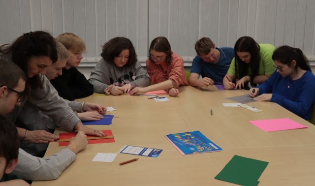 Na zdjęciu znajduję się młodzież siedząca przy złączonych stołach i rysują na kolorowych kartkach. W tle znajdują się okna. 