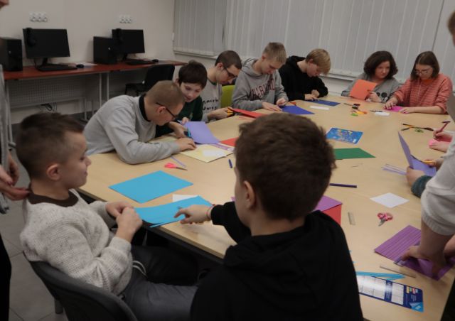 Na zdjęciu znajduję się młodzież iedząca w sali komputerowej przy złączonych stolikach i rysująca na kolorowych kartkach.