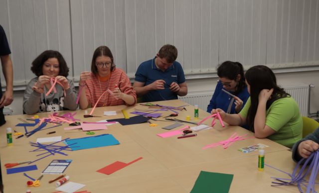 Na zdjęciu znajduję się piątka młodzieży siedząca przy złączonych stolikach i wycinająca paski z kolorowych kartek. W tle znajdują się okna.