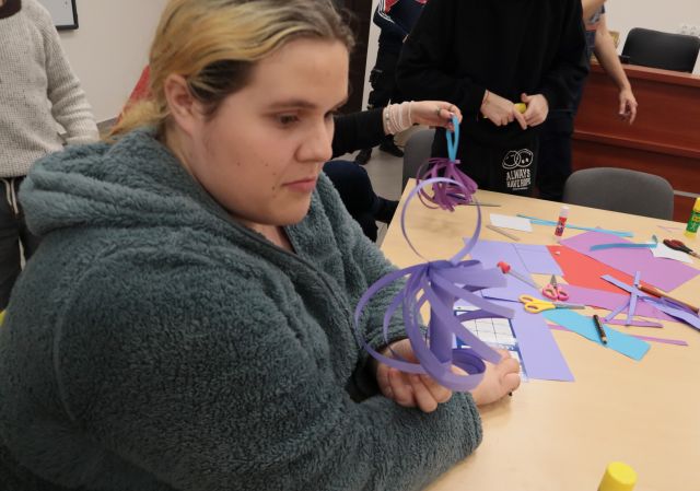 Na zdjęciu znajduję się nastolatka trzymająca w ręce zrobioną przez siebie fioletową papierową bombkę. W tle znajdują się kolorowe kartki na stoliku oraz inna skończona praca trzymana w ręce.
