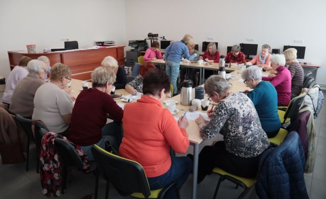 Na zdjęciu znajdują się seniorki siedzące i rozwiązujące zadania przy stolikach złączonych w podkowę, na których znajdują się termosy i kubki. Znajdują się w sali komputerowej.