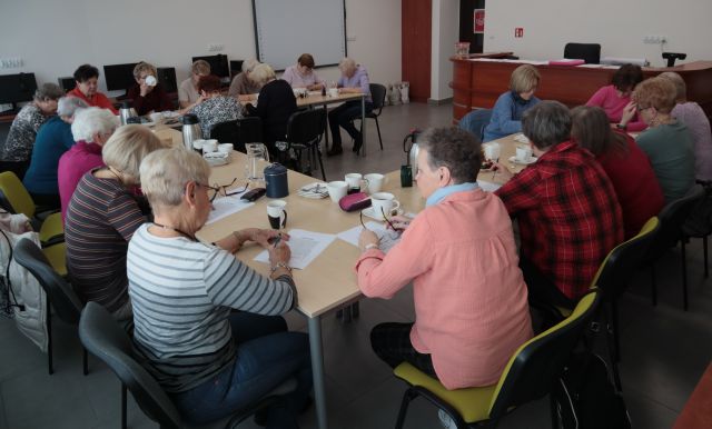 Na zdjęciu znajdują się kobiety siedzące i rozwiązujące zadania przy stołach złączonych w podkowę, na których znajdują się kartki oraz ciepłe napoje. Znajdują się w sali komputerowej.