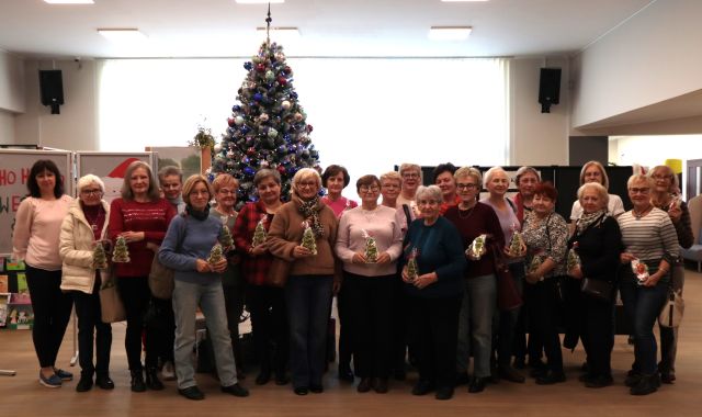 Na zdjęciu znajdują się dwadzieścia dwie kobiety stojące na tle choinki i trzymające w dłoniach pierniczki w kształcie choinki. W tle znajdują się okna.