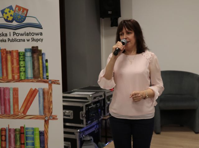Na zdjęciu znajduję się kobieta w różowej bluzce z mikrofonem przy ustach. W tle znajduję się fotel oraz baner z książkami oraz logiem biblioteki.