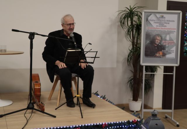 Na zdjęciu znajduję się mężczyzna, Krzysztof Daukszewicz siedzący na krześle na scenie, przed nim stoi mikrofon na statywie oraz statyw do nut, a obok niego leży gitara. Przy scenie znajduję się duży kwiat oraz plakat.