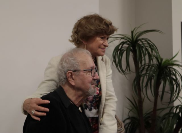 Na zdjęciu znajduję się mężczyzna oraz stojąca obok niego kobieta, która trzyma rękę na jego ramieniu. Są widoczni od pasa w górę, a w tle po prawej stronie znajduję roślina. Zdjęcie zrobione od boku.
