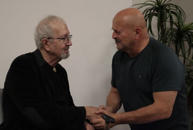 Na zdjęciu widać siedzącego mężczyzna i stojącego przed nim innego mężczyznę, podają sobie rękę i rozmawiają.