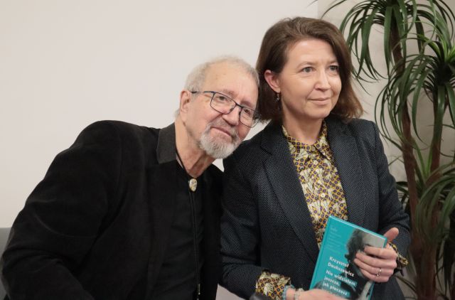 Na zdjęciu znajduję się mężczyzna i kobieta trzymająca książkę, mężczyzna lekko przechyla się w stronę kobiety. Po prawej stronie znajduję się zielona roślina.