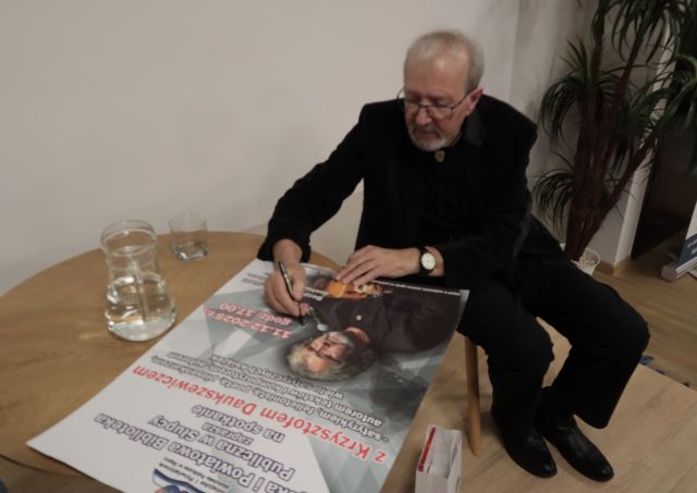 Na zdjęciu znajduję się siedzący przy stole mężczyzna, który podpisuje plakat.